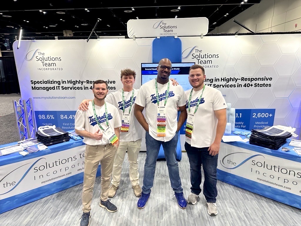 Four members of The Solutions Team stand smiling at their trade show booth featuring banners about managed IT services across 40+ states.